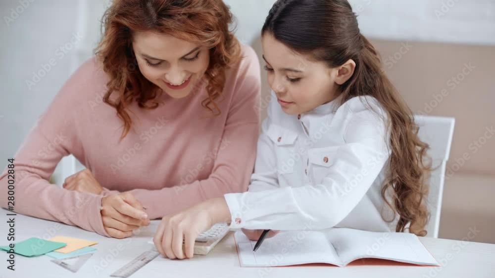 smiling preteen schoolgirl turning pages in copy book and talking to mother while she typing on calculator smiling preteen schoolgirl turning pages in copy book and talking to mother while she typing on calculator