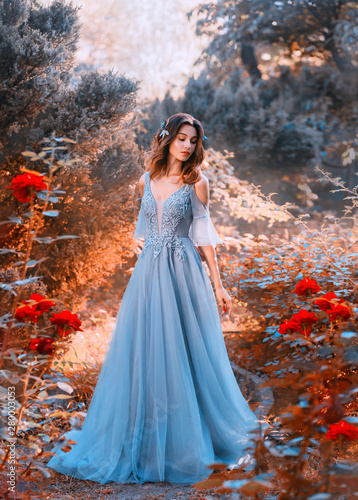 sad princess walks in fading autumn garden with withered plants, lady with short dark hair in chic light blue sky dress looks at red roses with sadness, clean young attractive girl, creative colors.