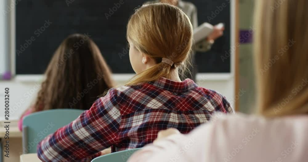 Rear of the schoolgirl sitting in the classroom during lesson ad her friend, cute girl, turning to her from the desk in front and whispering while laughing. Close up. Back view. Rear of the schoolgirl sitting in the classroom during lesson ad her friend, cute girl, turning to her from the desk in front and whispering while laughing. Close up. Back view.