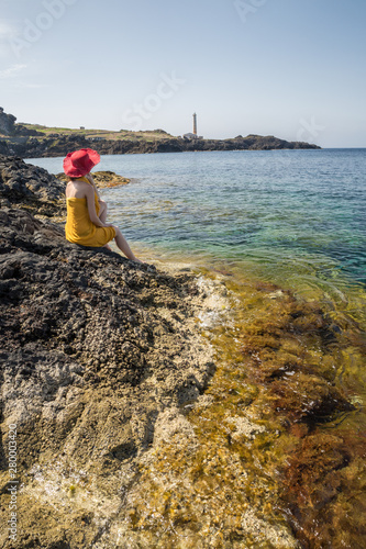 Ustica island, Sicily. Woman sitting on a rock looking at the sea. Summer holidays. Rocky coast, calm sea and transparent blue water 