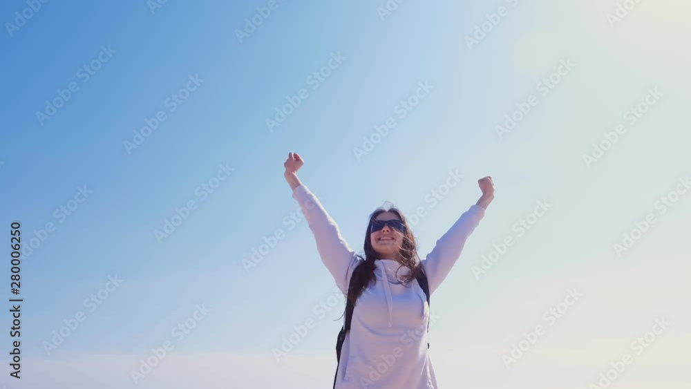 Happy girl winner in sunglasses with backpack stands in victory pose with raising hands on blue sky background. Travelling and winner concept. Carefree young woman on vacation. Happy girl winner in sunglasses with backpack stands in victory pose with raising hands on blue sky background. Travelling and winner concept. Carefree young woman on vacation.