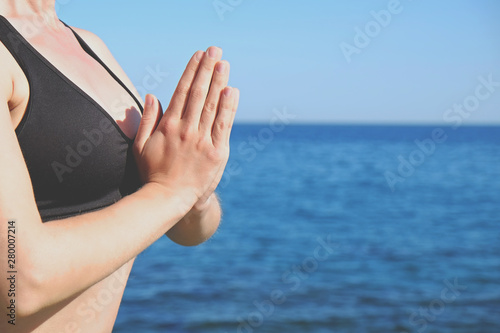 Cropped shot of attractive sporty young yogi woman wearing sportswear practicing yoga, sitting with namaste gesture, meditating on cliff over the calm ocean background. Close up, copy space.