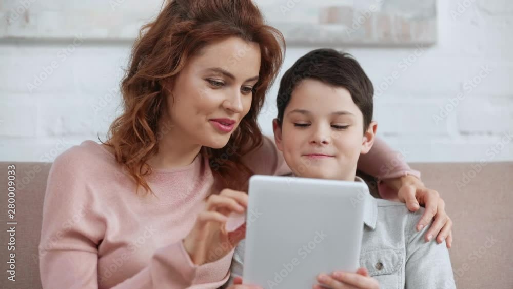 smiling mother embracing son and using digital tablet while sitting on sofa in living room smiling mother embracing son and using digital tablet while sitting on sofa in living room