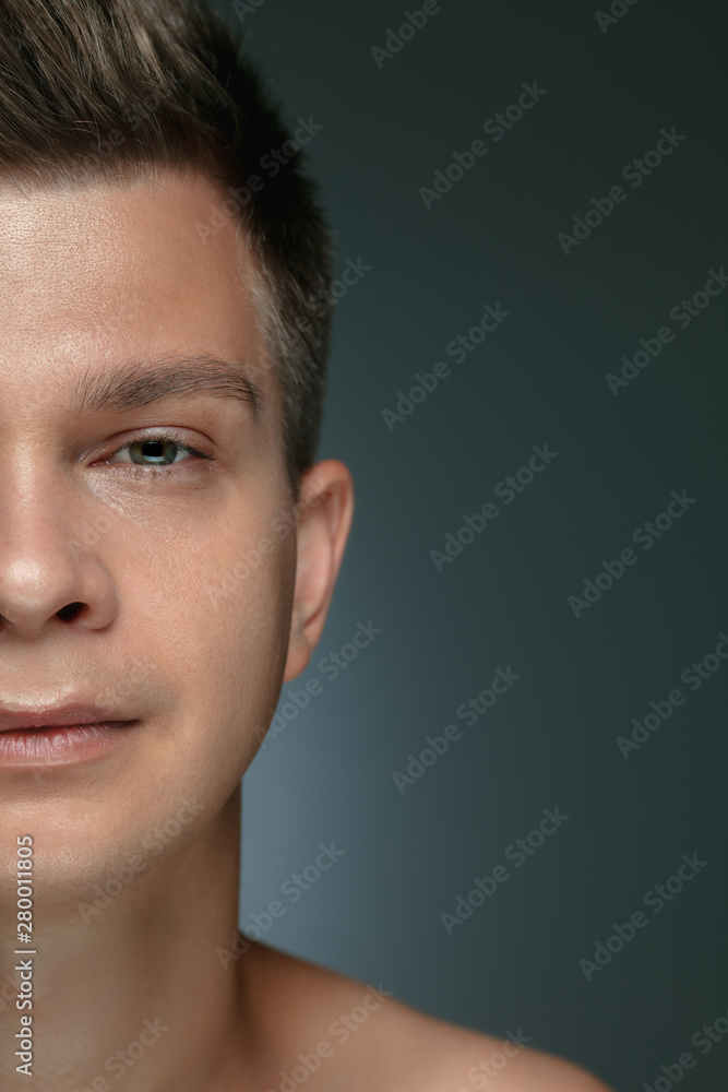 Close-up portrait of young man isolated on grey studio background. Caucasian male model looking at camera and posing, smiling. Concept of men's health and beauty, self-care, body and skin care.