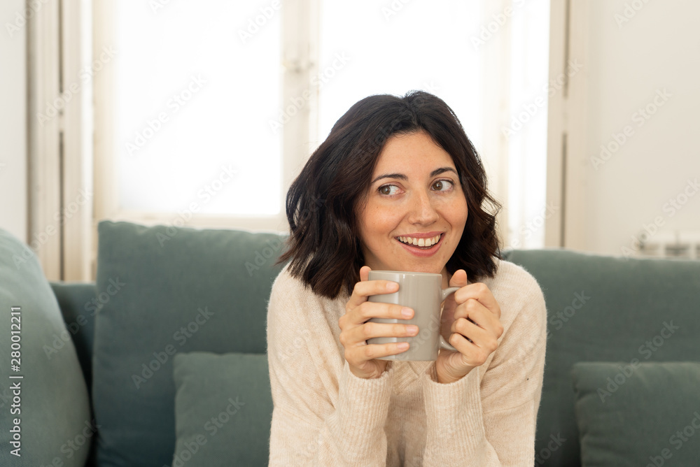 Happy young woman sitting on sofa at home with a hot beverage. In leisure and spare time concept Happy young woman sitting on sofa at home with a hot beverage. In leisure and spare time concept
