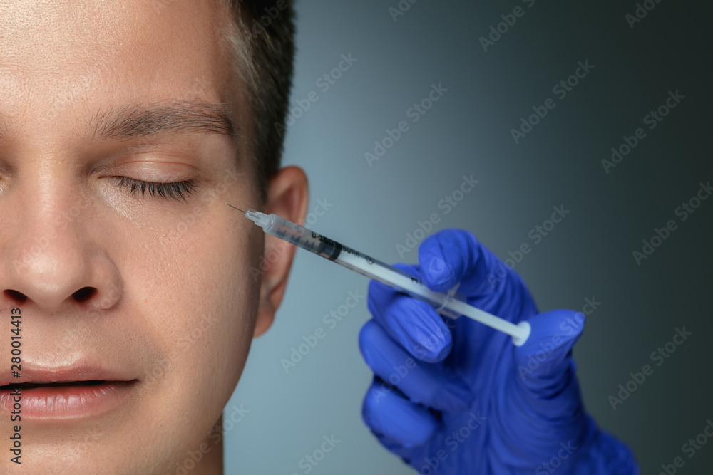 Close-up portrait of young man isolated on grey studio background. Filling surgery procedure. Concept of men's health and beauty, cosmetology, self-care, body and skin care. Anti-aging. Close-up portrait of young man isolated on grey studio background. Filling surgery procedure. Concept of men's health and beauty, cosmetology, self-care, body and skin care. Anti-aging.