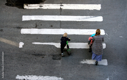 A woman with a stroller and her son on a bicycle crossing the road on a spring day. Back view from the window © Maria