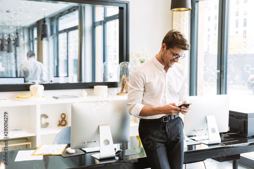 Image of masculine businesslike man holding cellphone while working in office
