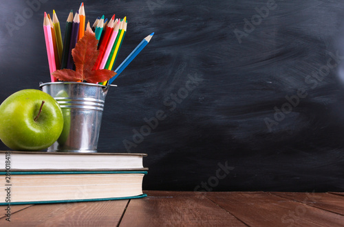Stack of books, green apple, colored pencils and autumn leaf on wooden desk. Classroom blackboard with copy space in background. Education, back to school concept