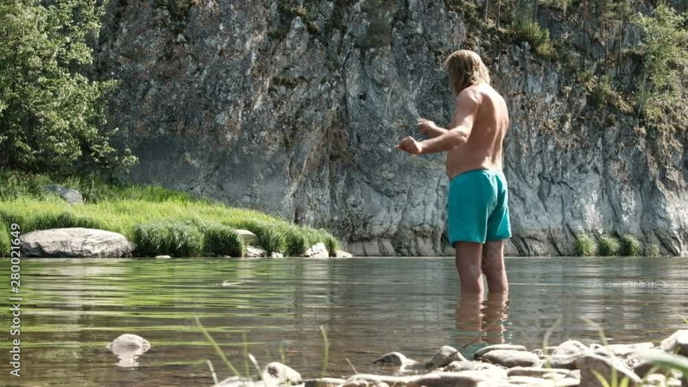 Fly fishing. An elderly fisherman stands knee-deep in the water of a mountain river. A bare man in blue shorts on the nature of fishing. Sport or hobby. Santa Claus in the summer on vacation. Fly fishing. An elderly fisherman stands knee-deep in the water of a mountain river. A bare man in blue shorts on the nature of fishing. Sport or hobby. Santa Claus in the summer on vacation.