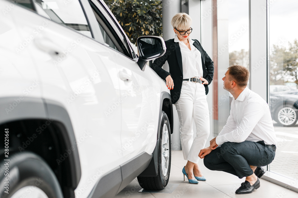 Wife and husband choose a car in dealership. Woman leaned on the car and man sits next it Wife and husband choose a car in dealership. Woman leaned on the car and man sits next it