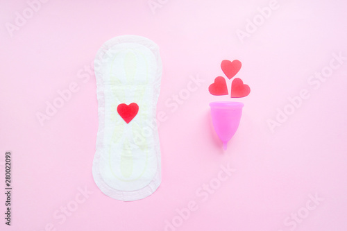 Reusable menstrual cup with red hearts and menstrual pad on pink background, Concept female intimate hygiene period products and zero waste. Flat lay, minimalism, top view. copyspace