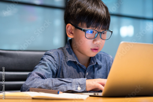 Smart looking Asian preteen boy writing and using computer laptop to study online lessons, research for project from home during Covid-19 pandemic, city lockdown and social distancing. photo