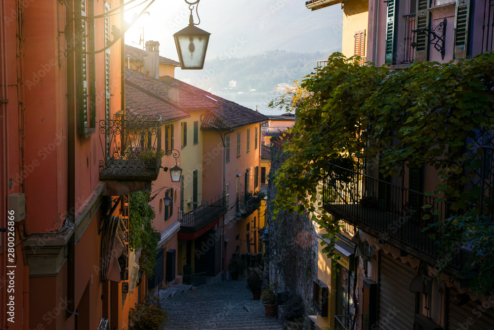 Narrow street with beautiful colourful houses with lantern and balcony in scenic Bellagio, Italy, region of Lombardy. The stairs down the Lake Como shore, mountains on the background. 