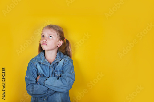 Little funny girl dressed in blue jeans shirt standing with arms folded on yellow background.  Children emotions and expressions concept photo