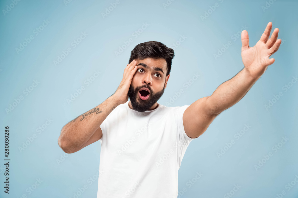 Fototapeta premium Half-length close up portrait of young hindoo man in white shirt on blue background. Human emotions, facial expression, sales, ad concept. Negative space. Pointing up being shocked and astonished.
