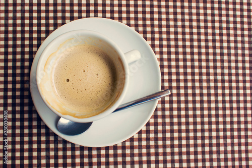 Coffee cup standing on a saucer with teaspoon on the table covered with checkered material. Top view. 