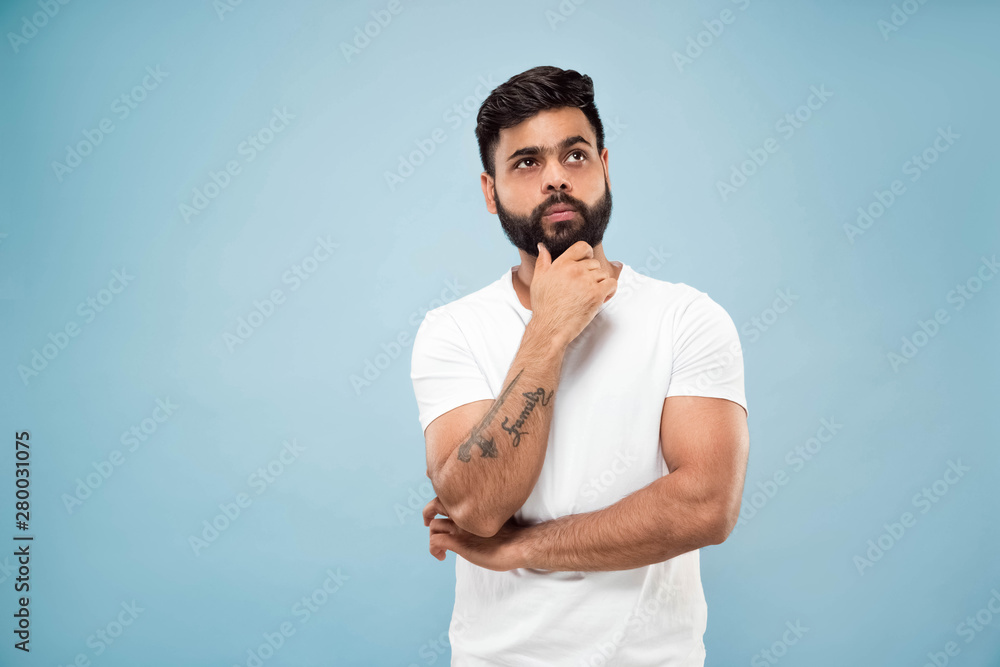 Half-length close up portrait of young hindoo man in white shirt on blue background. Human emotions, facial expression, ad concept. Negative space. Thinking while holding hand on his beard. Choosing.