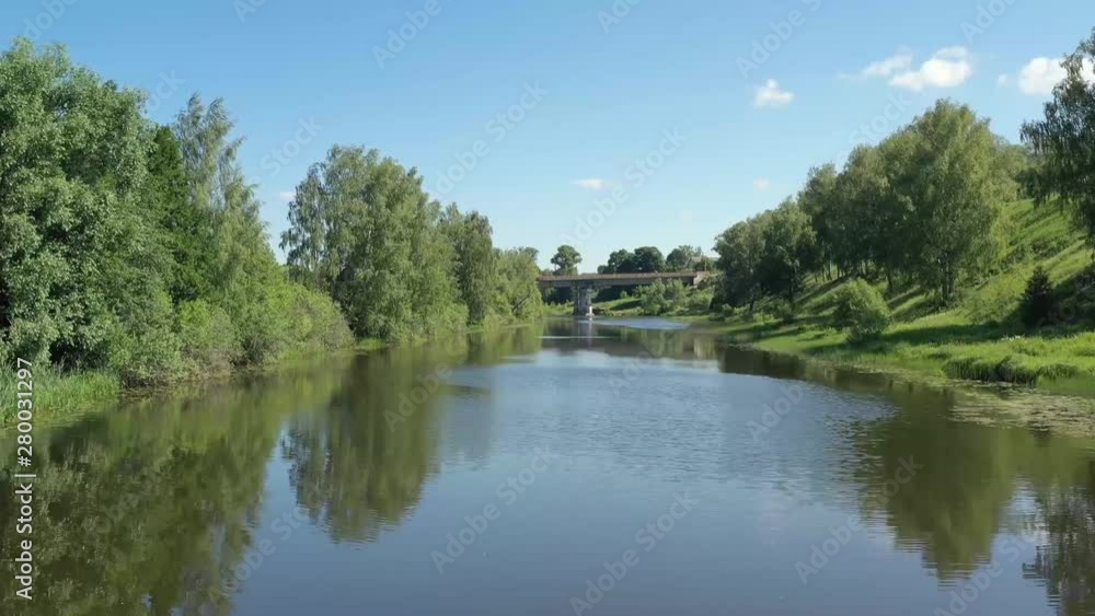 A quadcopter span along a beautiful, calm river on a sunny summer day with a road bridge in front A quadcopter span along a beautiful, calm river on a sunny summer day with a road bridge in front