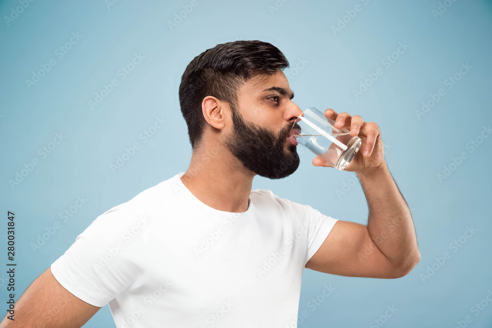 Half-length close up portrait of young hindoo man in white shirt on blue background. Human emotions, facial expression, ad concept. Negative space. Drinking pure water, looks calm and happy. Half-length close up portrait of young hindoo man in white shirt on blue background. Human emotions, facial expression, ad concept. Negative space. Drinking pure water, looks calm and happy.