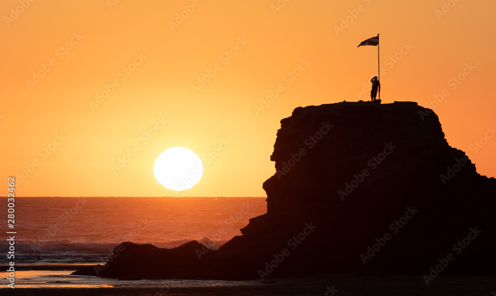 Sunset on the Atlantic Ocean in Cornwall in Perranporth. A man stands on a rock under a flag and looks into the setting sun. Very romantic.