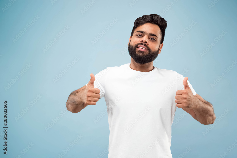 Half-length close up portrait of young hindoo man in white shirt on blue background. Human emotions, facial expression, ad concept. Negative space. Showing the sign of OK, nice, great. Smiling. Half-length close up portrait of young hindoo man in white shirt on blue background. Human emotions, facial expression, ad concept. Negative space. Showing the sign of OK, nice, great. Smiling.