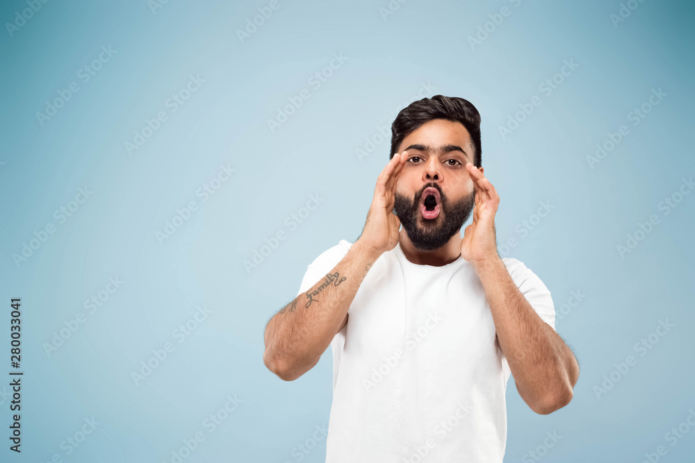 Half-length close up portrait of young hindoo man in white shirt on blue background. Human emotions, facial expression, sales, ad concept. Negative space. Calling for somebodies with hands on his face Half-length close up portrait of young hindoo man in white shirt on blue background. Human emotions, facial expression, sales, ad concept. Negative space. Calling for somebodies with hands on his face