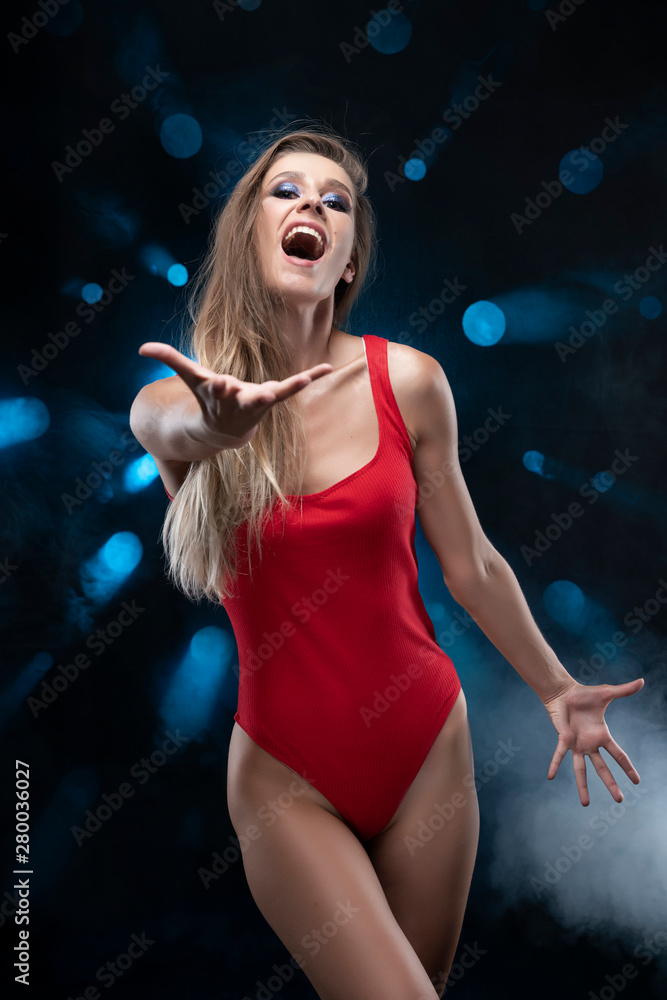 A beautiful long-haired slim girl wearing a red body, sings and screams and gives her hand against the background of spots of light from which blue rays shine in the smoke. Advertising and commercial.