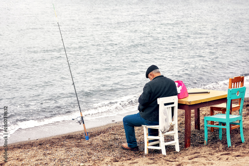 Old indigenous Turkish fisherman sitting on a wooden chair and fishing near the seaside on a cloudy winter day.