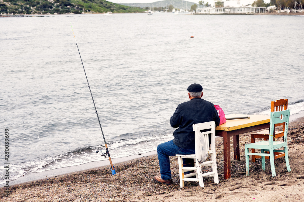 Old indigenous Turkish fisherman sitting on a wooden chair and fishing near the seaside on a cloudy winter day. Old indigenous Turkish fisherman sitting on a wooden chair and fishing near the seaside on a cloudy winter day.