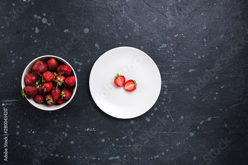 White ceramic bowl with ripe strawberries and a white plate with sliced ​​strawberries on the stone surface. Top view.