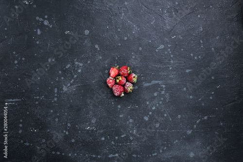 Strawberry berries in powdered sugar on a dark stone surface. Top view.