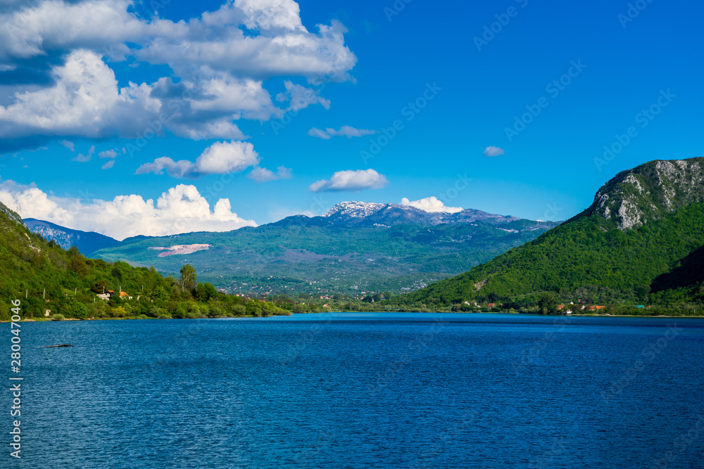 Montenegro, Barrier lake water of jezero liverovoci inside green valley near niksic city surrounded by green trees and forest covering rocky mountains and snow covered peaks under blue sky