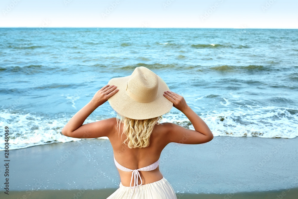Blonde Woman in White Summer Style Standing at Sea and Holding Hat. Luxury Lifestyle Rear View. Summer Vacation Concept