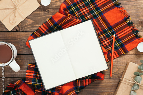 Autumn flatlay composition with red knitted scarf, blank book, cup of tea, craft envelope, pen, gift box on wooden background. Cozy autumn or winter holiday concept. Flat lay, top view, copy space.