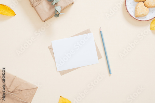 Autumn composition. Cozy feminine desktop with letter, blank paper card, gift box, cookies, fallen leaves on pastel beige background. Flat lay, top view, overhead. Autumn, fall concept.