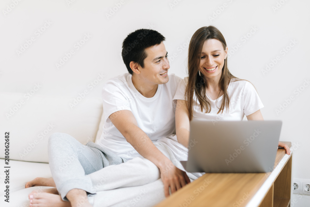 Portrait of happy young couple sitting on couch and working on laptop at home. Happy young couple is working on laptop in bright apartment. Portrait of happy young couple sitting on couch and working on laptop at home. Happy young couple is working on laptop in bright apartment.