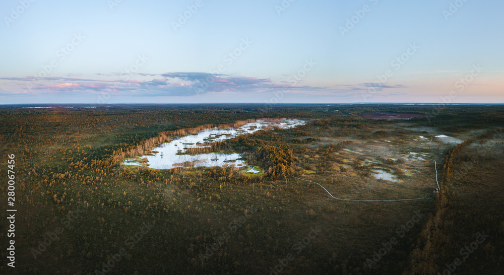 Warmly colored sunrise over a foggy swamp. Aerial view of stunning landscape at peat bog at Cenas Tirelis in Latvia. Wooden trail leading along the lake surrounded by pounds and forest.
