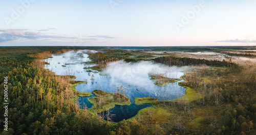  Warmly colored sunrise over a foggy swamp. Aerial view of stunning landscape at peat bog at Cenas Tirelis in Latvia. Wooden trail leading along the lake surrounded by pounds and forest.  © Viesturs
