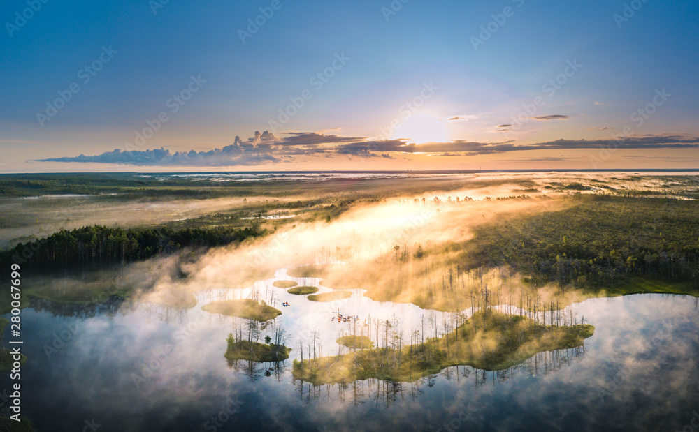 Obraz premium Warmly colored sunrise over a foggy swamp. Aerial view of stunning landscape at peat bog at Cenas Tirelis in Latvia. Wooden trail leading along the lake surrounded by pounds and forest.