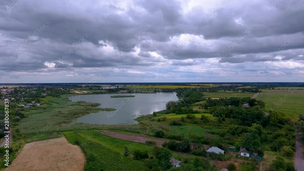 Ukrainian village on the lake with the eyes of a bird. Drone flies over the lake and nearby are private houses in the village. Summer, cloudy weather. Ukrainian village on the lake with the eyes of a bird. Drone flies over the lake and nearby are private houses in the village. Summer, cloudy weather.