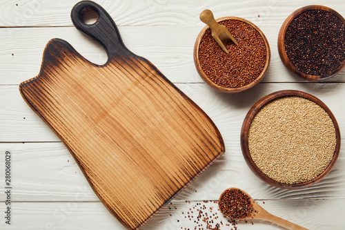 top view of white, black and red quinoa in wooden bowls near chopping board on white table