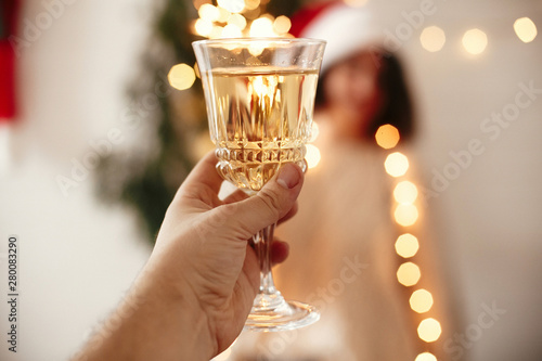 Man toasting with champagne glass to happy girl in santa hat with sparkler, celebrating at christmas tree lights in festive room. Happy New Year eve party. Happy Holidays
