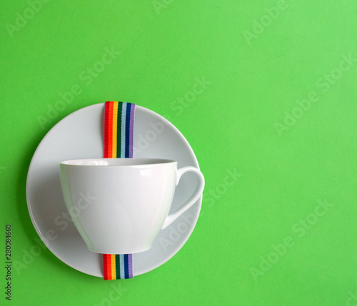White coffee Cup on green pastel colorful paper geometric flat background with rainbow ribbon lgbt on saucer. Drink Cup template for your design to place text, image and logo layout