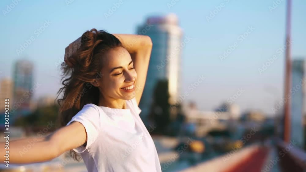 happy smiling young pretty woman in white t-shirt is standing on the bridge on blue sky background, hand straightens hair, enjoying peaceful sunset and looking at camera. calm and relaxation. slow happy smiling young pretty woman in white t-shirt is standing on the bridge on blue sky background, hand straightens hair, enjoying peaceful sunset and looking at camera. calm and relaxation. slow