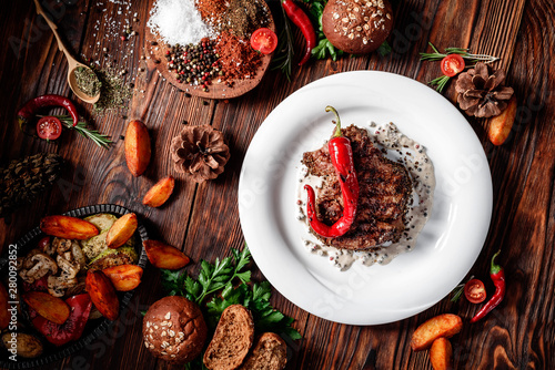 Juicy grilled steak decorated with chili peppers on a white plate, on dark wooden background in beautiful composition among vegetables and spices. Top view. Flat lay