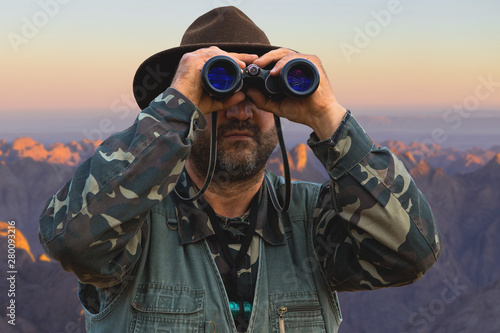 A hunter in a hat with binoculars looks out for prey.