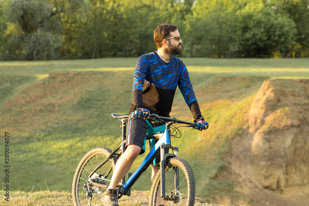 Cyclist in shorts and jersey on a modern carbon hardtail bike with an air suspension fork rides off-road on the orange-red hills at sunset evening in summer Cyclist in shorts and jersey on a modern carbon hardtail bike with an air suspension fork rides off-road on the orange-red hills at sunset evening in summer