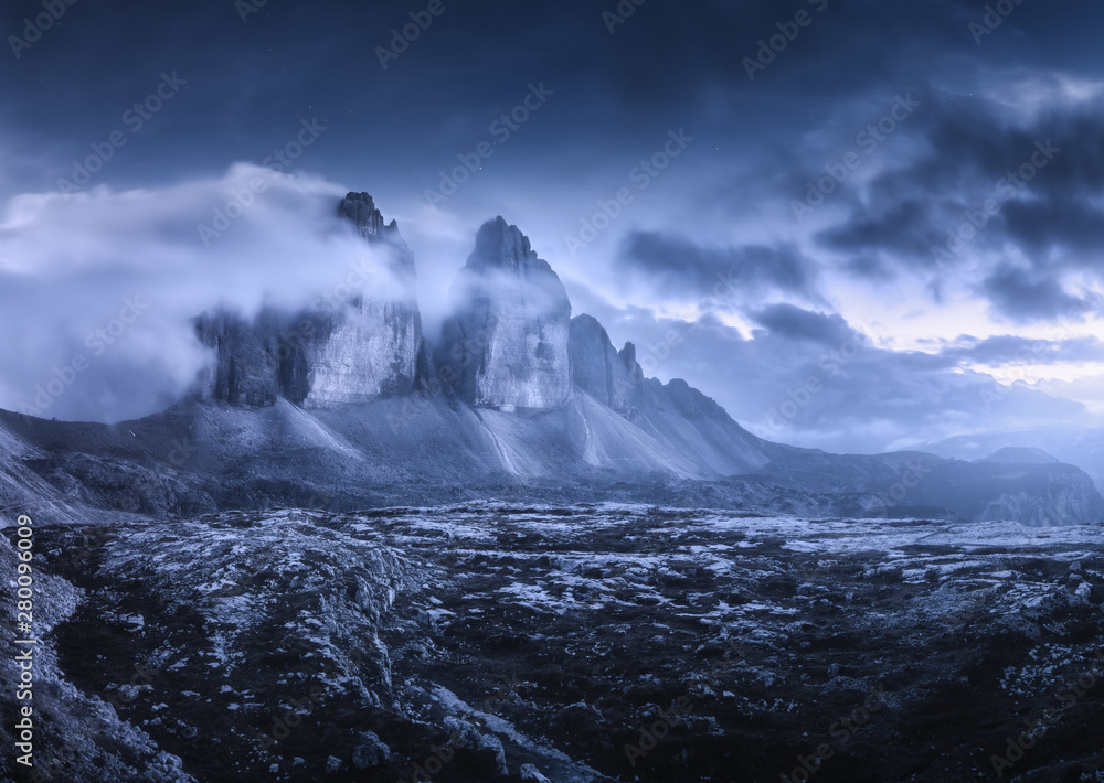 Mountains in fog at beautiful night. Dreamy landscape with mountain peaks, stones, grass, purple sky with blurred low clouds, stars and moon. Rocks at dusk. Tre Cime in Dolomites, Italy. Italian alps