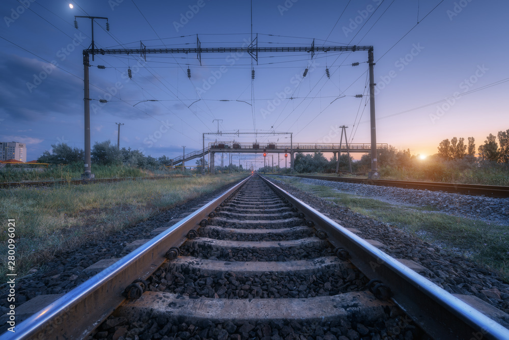 Railroad and blue sky with moon at sunset. Summer rural industrial landscape with railway station, sky with clouds and sunlight, green grass. Railway platform. Transportation. Heavy industry at dusk
